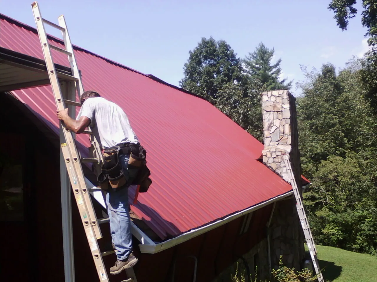 Roofer on a ladder beside a red metal roof and stone chimney — Soffit & Fascia Repair by Roofing Brothers in West Elkton
